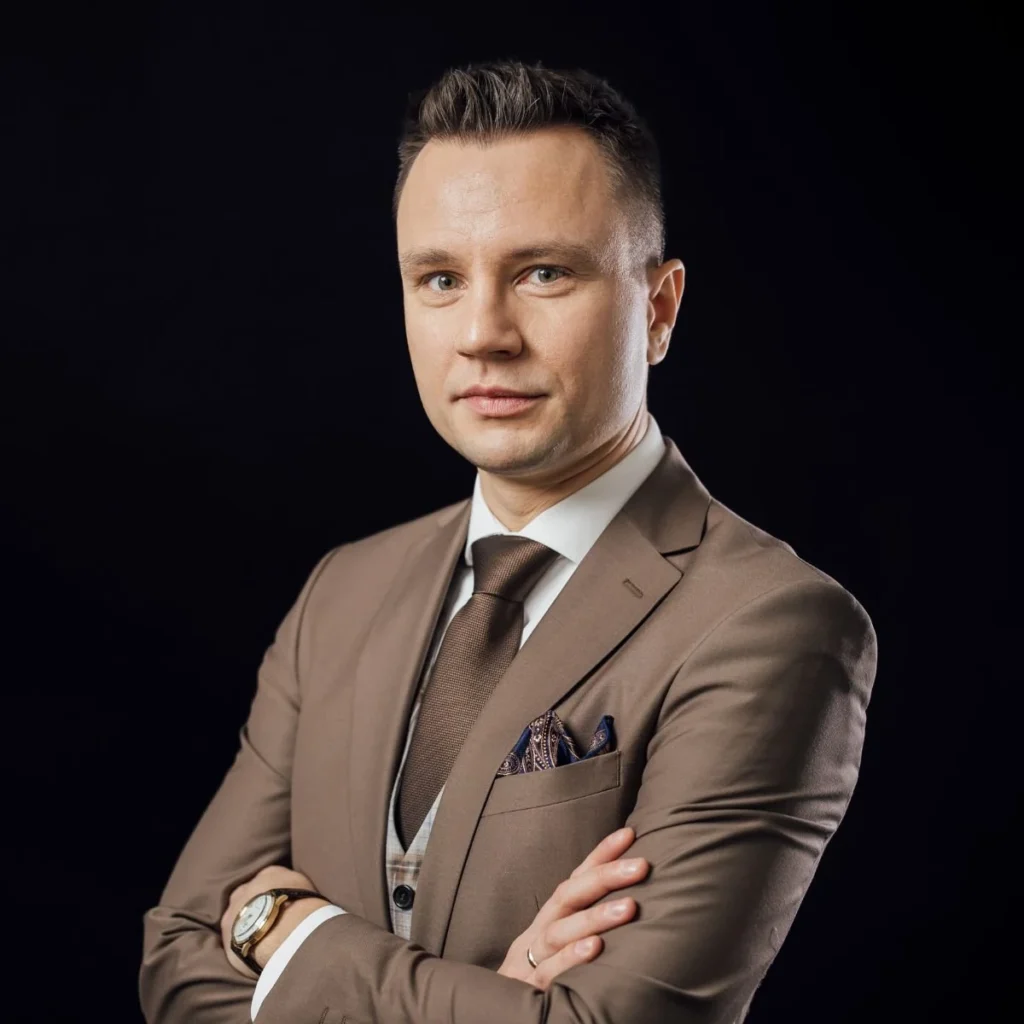 Piotr Sperka z założonymi rękami, w beżowym garniturze — portret na ciemnym tle