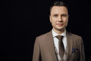 Piotr Sperka w brązowym garniturze z kamizelką — portret poziomy na ciemnym tle